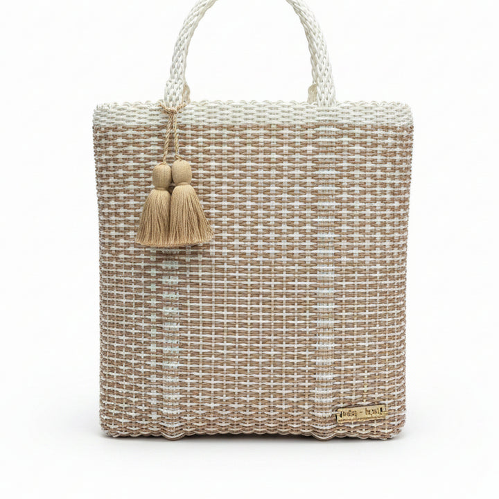 A woven tote bag made from natural beige and off-white raffia or straw fibers, featuring two braided top handles folded over and a beige tassel charm on the left side. A small wooden brand tag is visible in the bottom right corner.