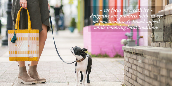 Person holding large cesta tote in traditional design of white & sunflower. Small dog on leash  looking at person. colorful street scape in Takoma Park. Text on image says 'we focus on creativity ~ designing our products with an eye on practical use & broad appeal while honoring local traditions'